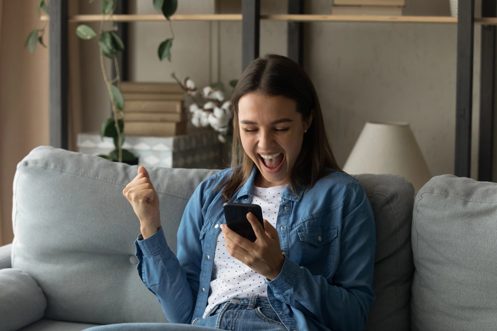 A woman sitting on a couch, cheering with excitement while looking at her phone.