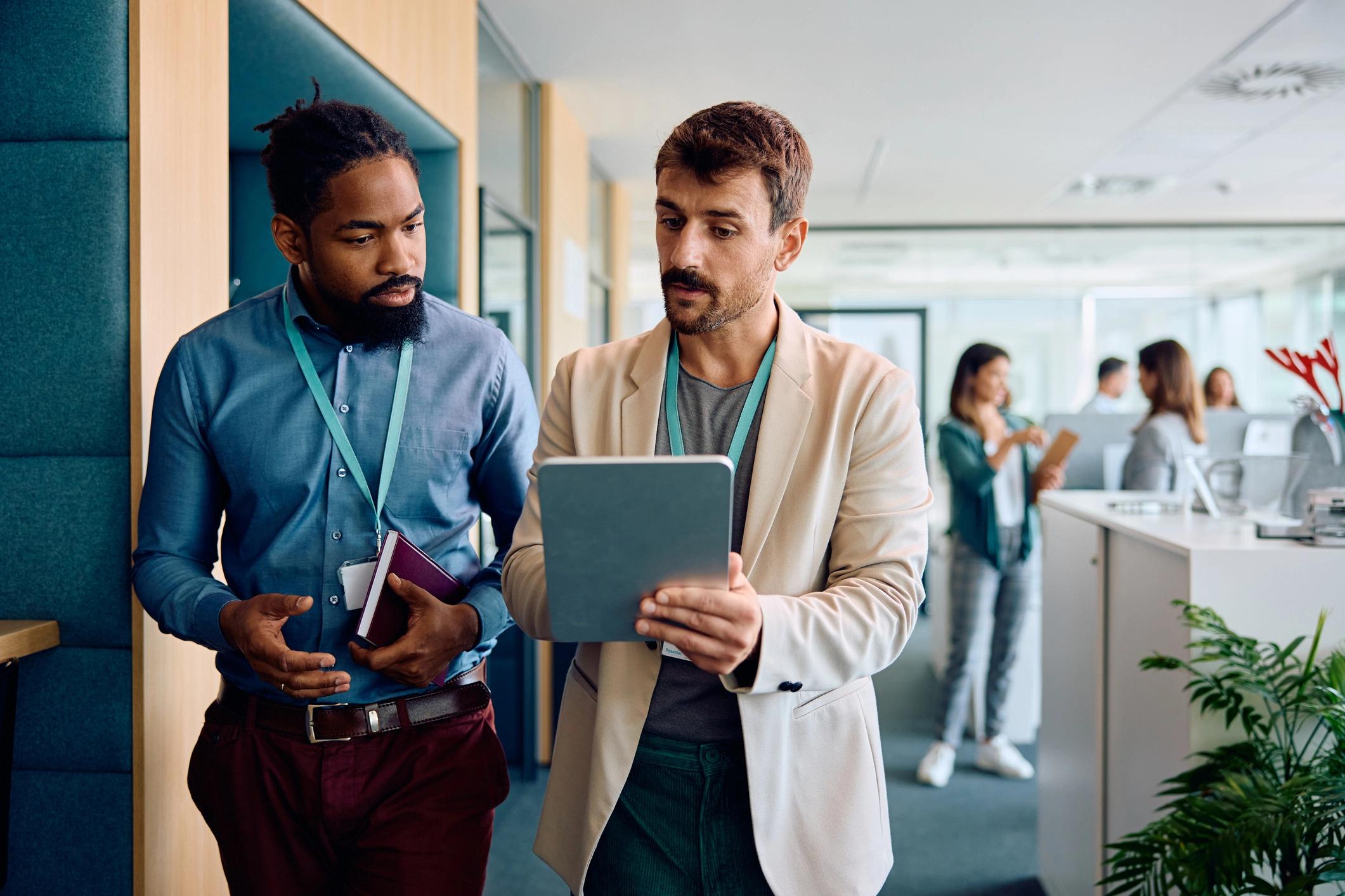 Two men in business attire having a discussion while looking at a tablet in an office setting.