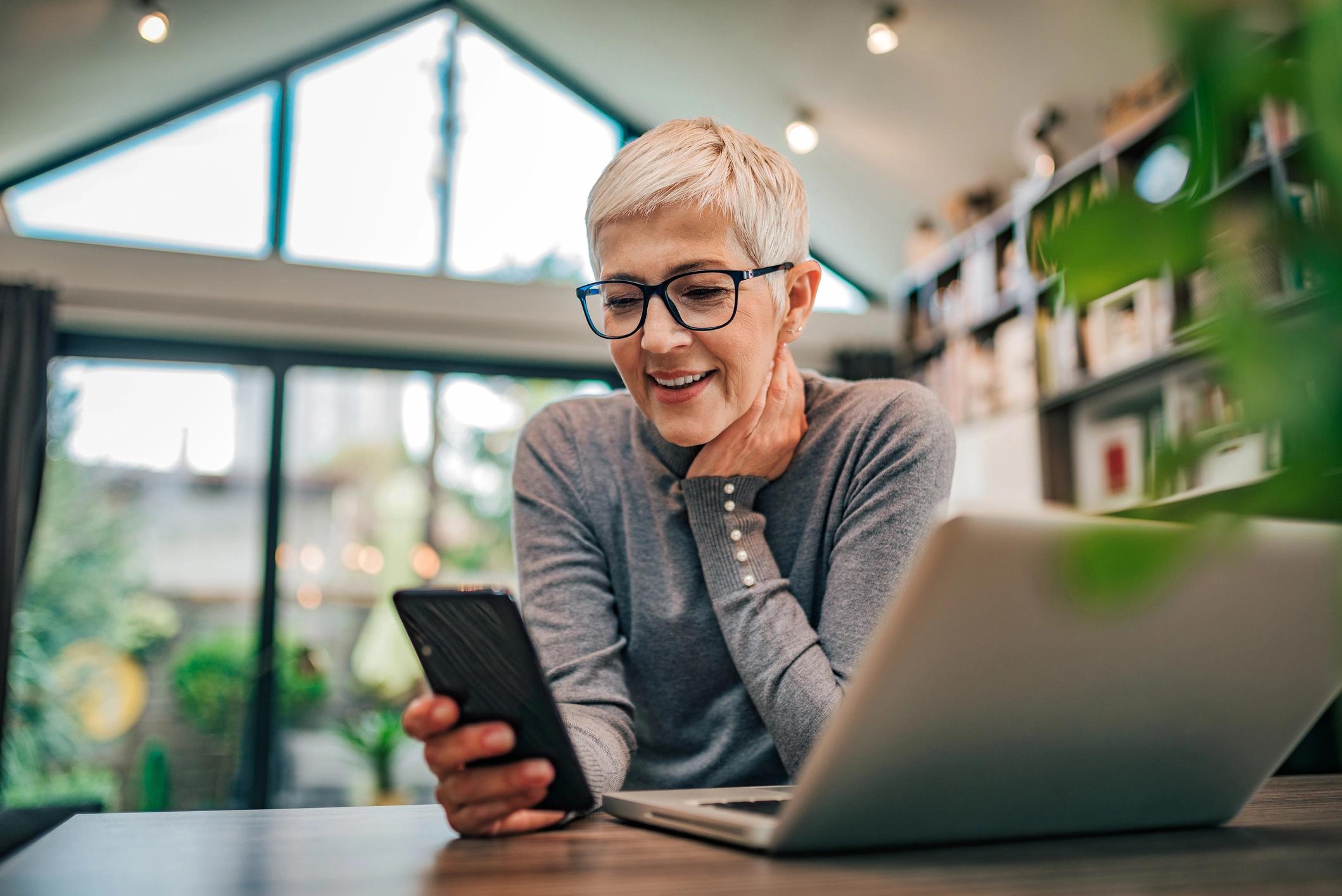 An older woman with glasses smiles while looking at her phone, sitting at a table with a laptop in a bright room.