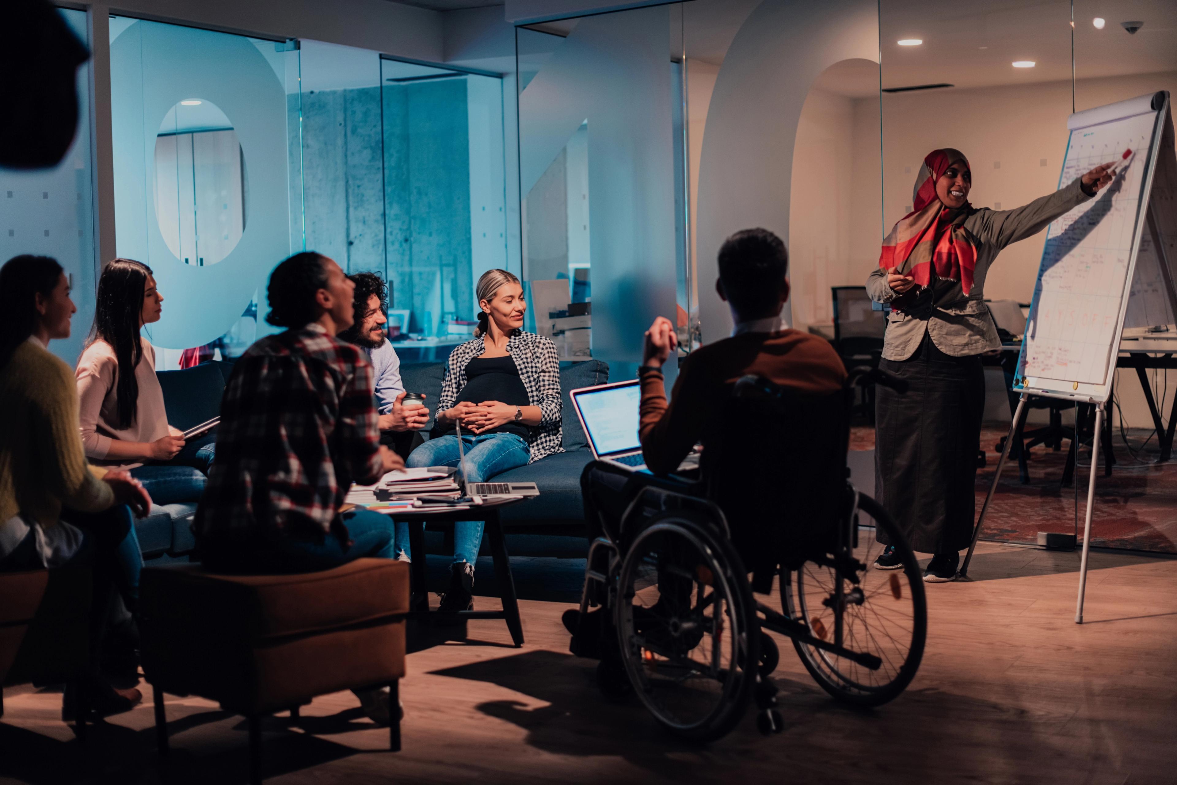 A diverse group of people in a meeting, including a person in a wheelchair and a woman in a hijab presenting.