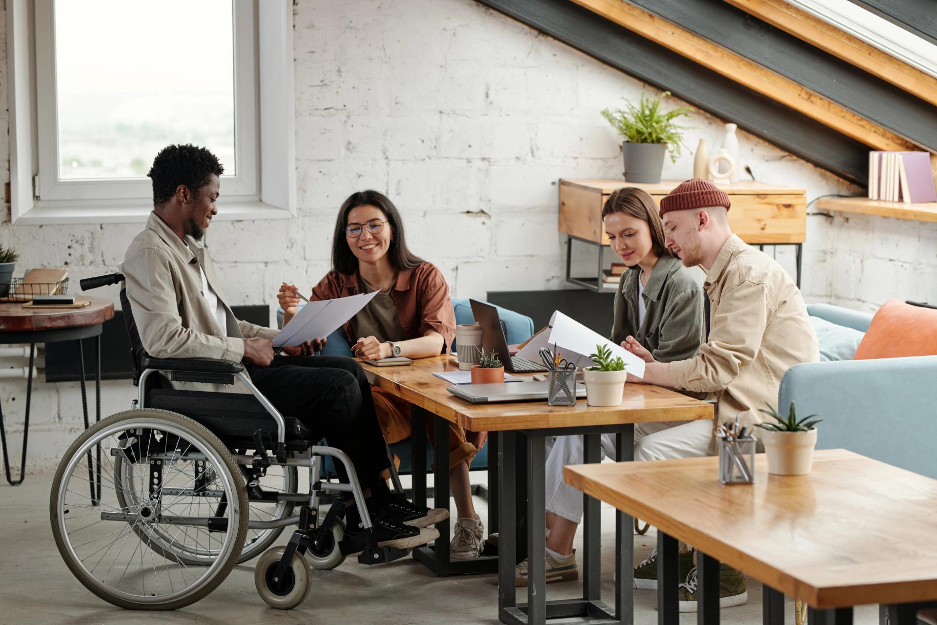 A group of people, including a person in a wheelchair, collaborating around a table in a bright office.