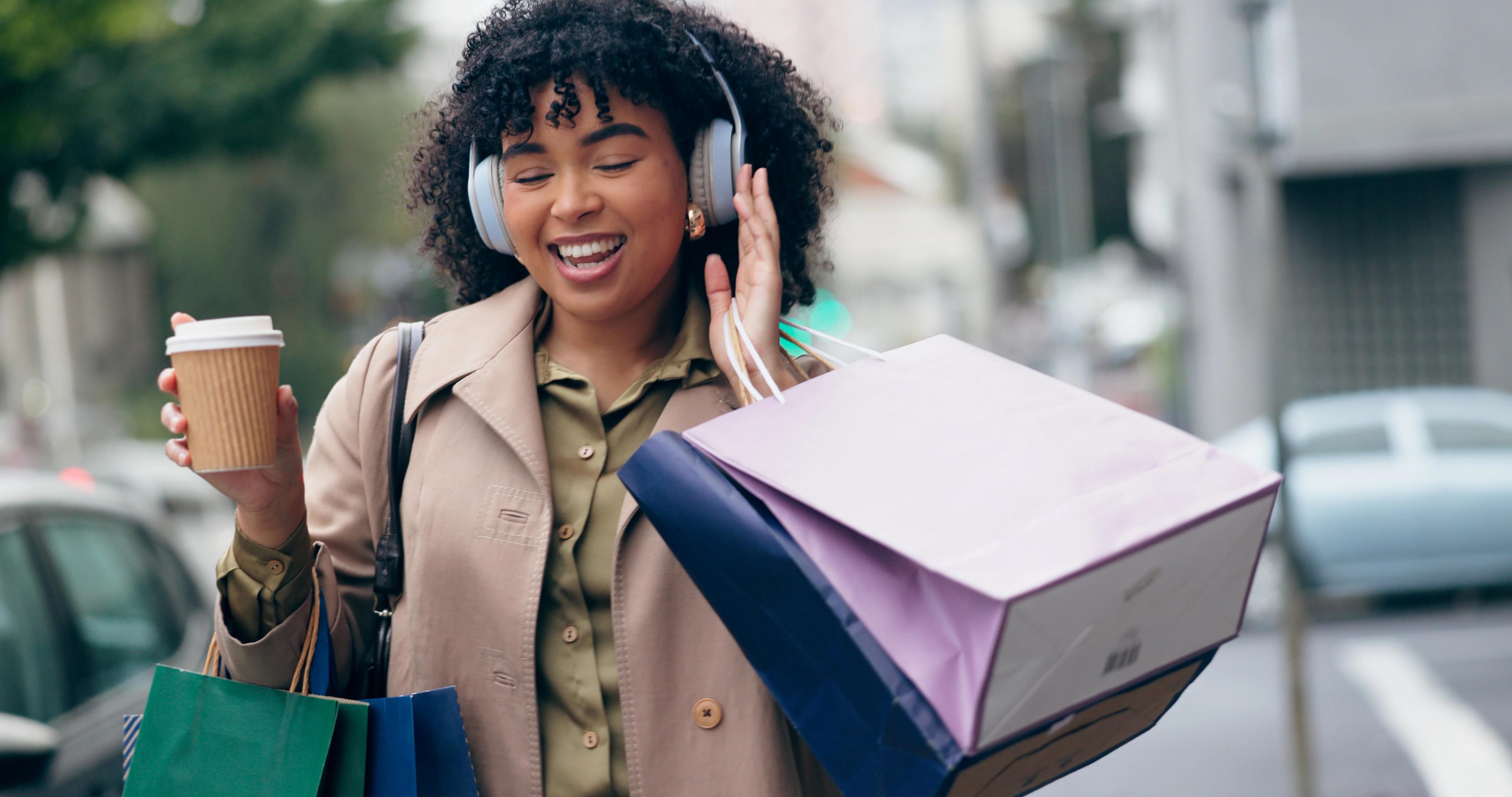 A woman enjoying shopping, holding bags and a coffee cup, smiling while listening to music with headphones.