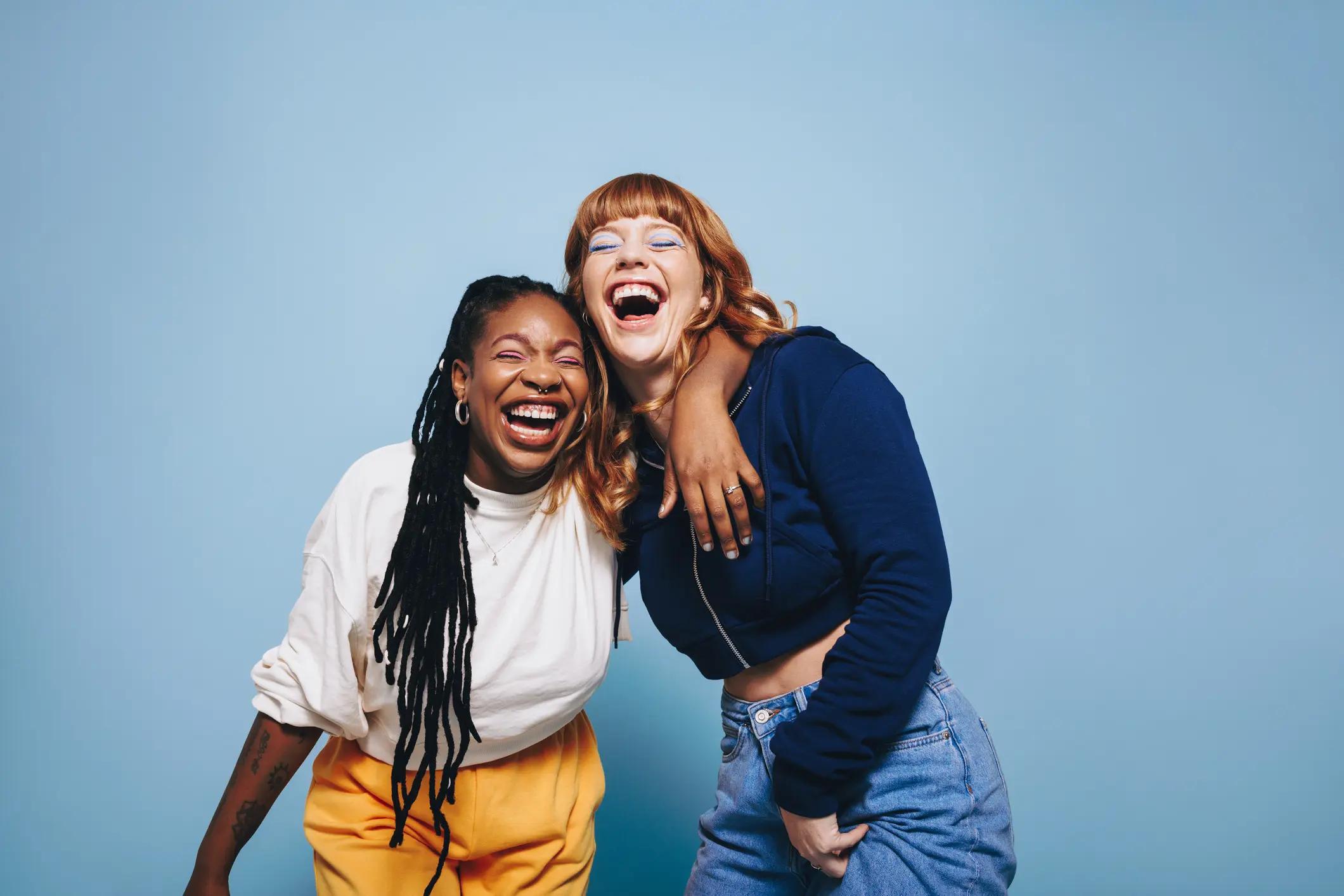 Two women are laughing together against a light blue background, looking bright and smiling.