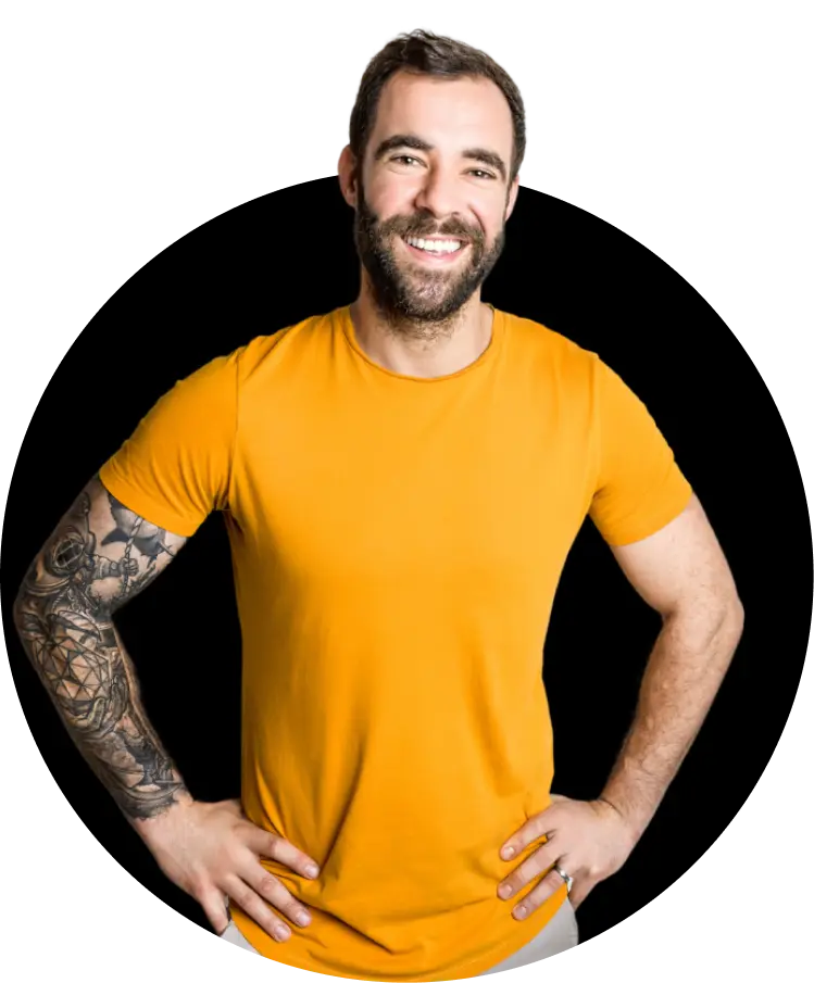 A man with light skin and a beard, wearing a yellow T-shirt. He has tattoos on his arms and is smiling broadly with his hands on his hips.