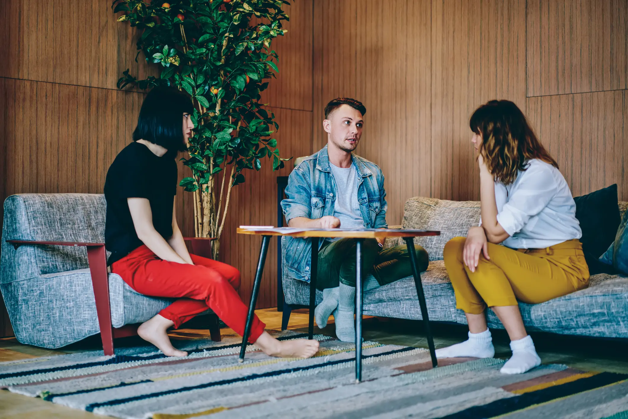 Three people are chatting in a cozy room. A man in a denim jacket sits on a couch, a woman in a white blouse and yellow pants sits on another couch, and a woman in a black top and red pants sits on an armchair.